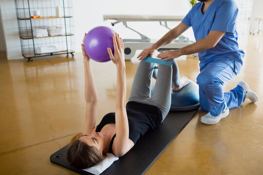 Female patient lying down on a mat, exercising with her physiotherapist using resistance band for legs and a fit ball, in a gymnasium or clinic. Physical therapy concept. physiotherapy